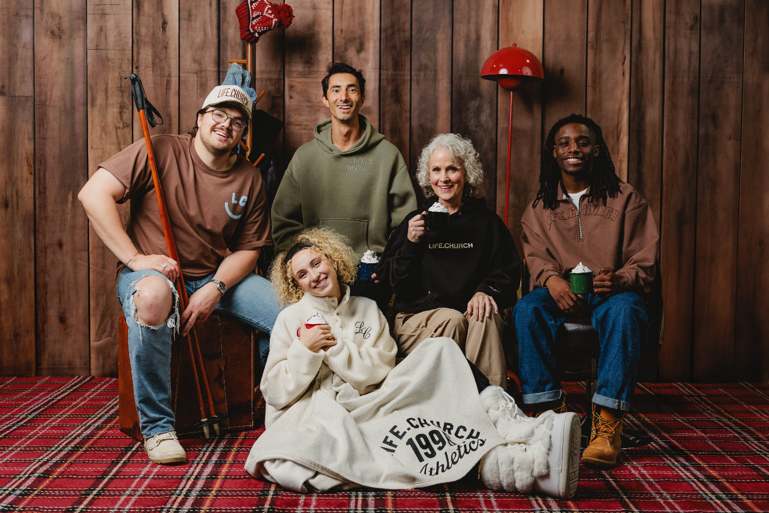 Five people posing together in a cozy indoor setting with a wood-paneled wall, red plaid rug, and holiday decorations. 