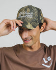 A man wearing a camouflage baseball cap with white embroidered text that reads 'Life.Church ALL DEVOTED FOLLOWER OF CHRIST' and adjusting the brim, against a plain light gray background.