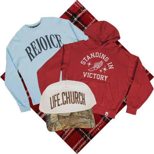 A flat-lay product bundle featuring a light blue sweatshirt with 'REJOICE' in dark blue text, a red hoodie with 'STANDING IN VICTORY' and a wing graphic in white, a white cap with 'LIFE.CHURCH' in brown text over a camouflage brim, and a red plaid blanket on a white background.