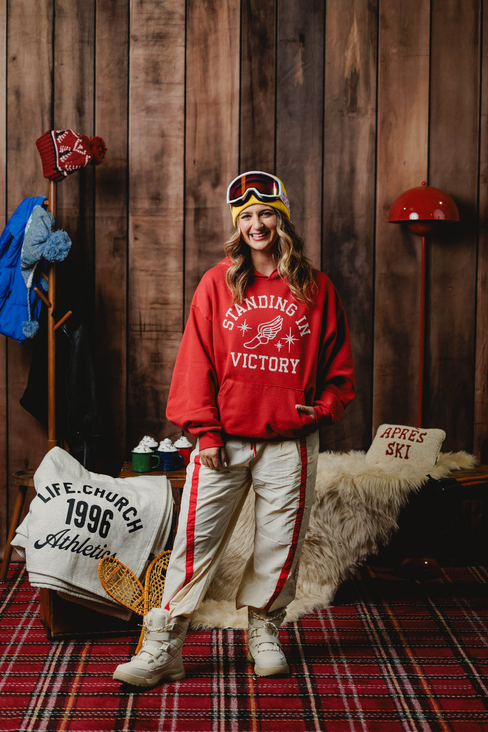 A woman stands in a rustic wood-paneled room wearing a red sweatshirt with 'STANDING IN VICTORY' printed on the front, cream ski pants with red stripes, a yellow beanie, and ski goggles. A red plaid rug covers the floor, and winter-themed decor, including a coat rack and a faux-fur rug, surrounds her.